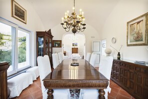 A dining room with a large wooden table, white upholstered chairs, a chandelier, and a view through large windows. - Suite Villa Carolina (Capri)