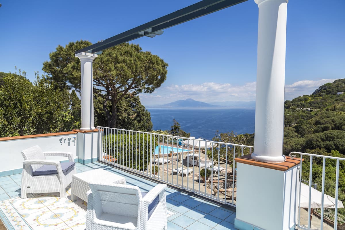 A balcony with white wicker furniture, a pool, and a scenic view of the sea and mountains.