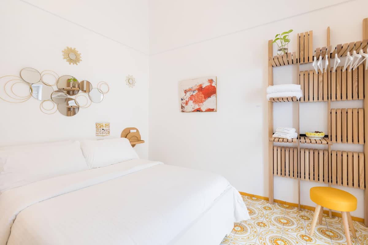 A hotel room with a bed, a wooden shelf with towels, a small stool, and wall decor.