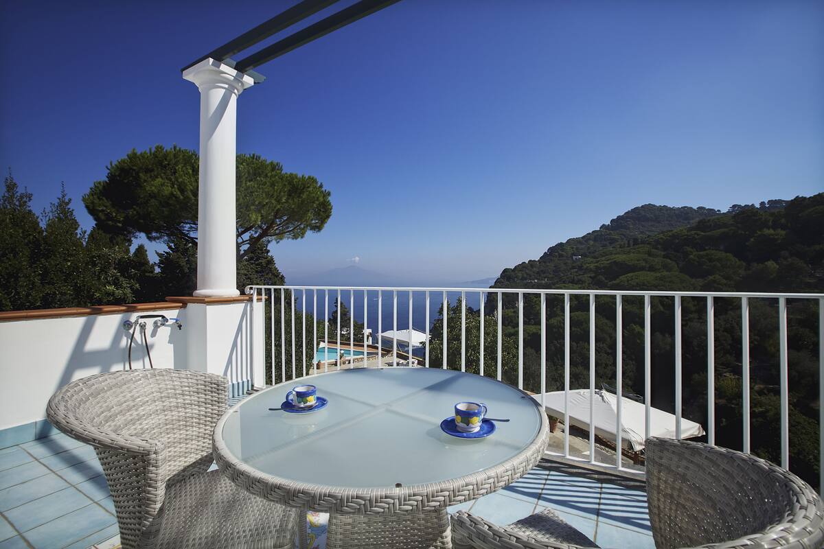 A round glass table with wicker chairs and two blue cups on a balcony with a white railing, overlooking a mountainous landscape.