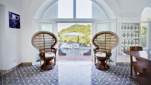 Una habitación con un amplio ventanal de arco, sillas de mimbre, una mesa de comedor y vistas a un jardín exuberante.