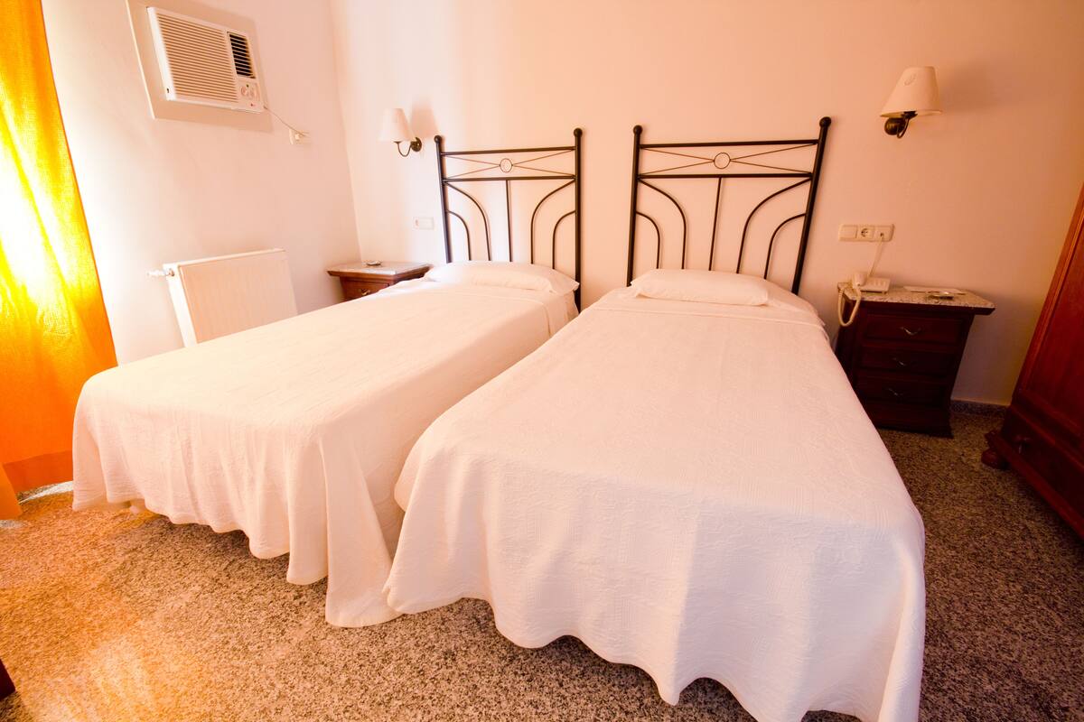 Two single beds with metal frames, a bedside table, a nightstand, a lamp, and a wall-mounted air conditioner.