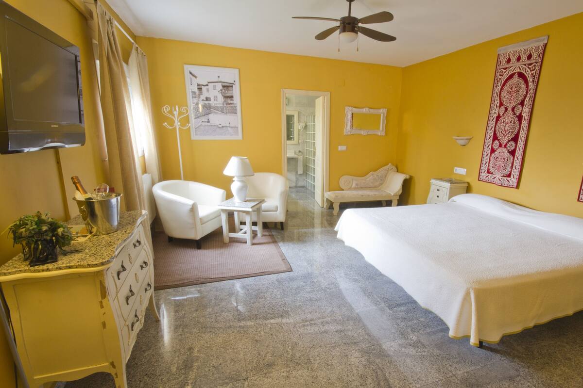 A hotel room with a bed, a chair, a small table, a TV, a ceiling fan, and a bathroom door.