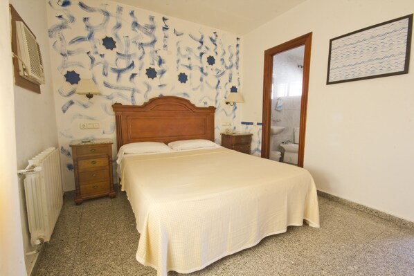 Un dormitorio con una cama, cabecera de madera, dos mesitas de noche, un radiador y una pared con un estampado azul y blanco.