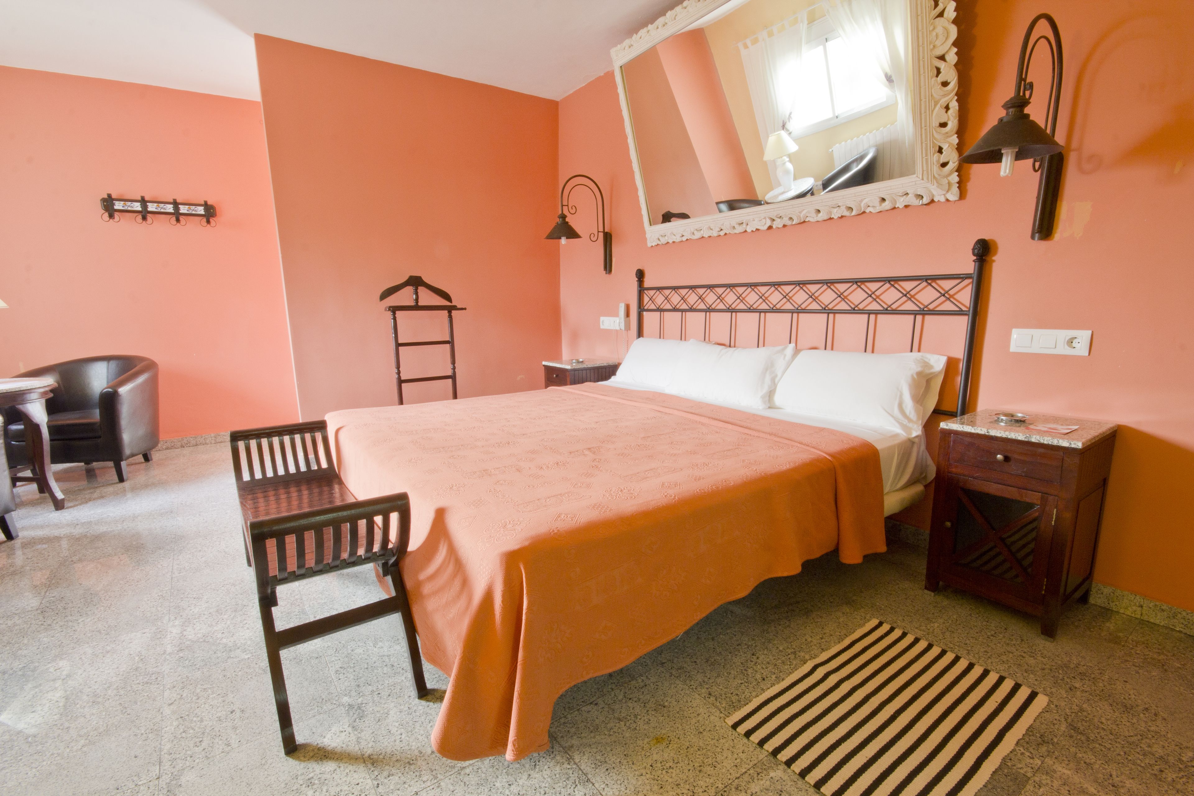 A bedroom with a bed, a nightstand, a chair, a wooden bench, a mirror, and a wall-mounted lamp.