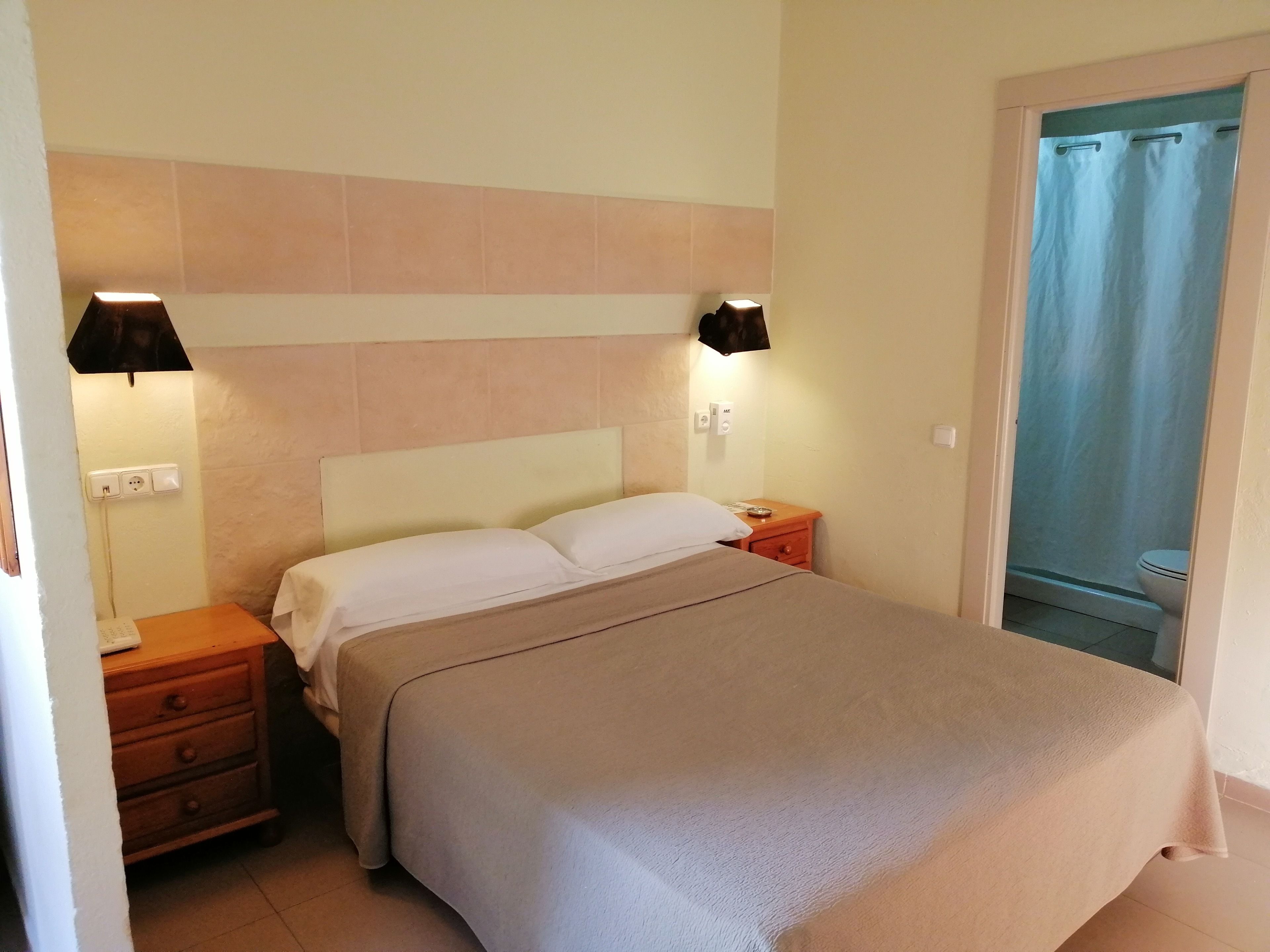 Una cama bien hecha con una colcha color beige, una mesita de noche con una lámpara y un aplique en la pared.