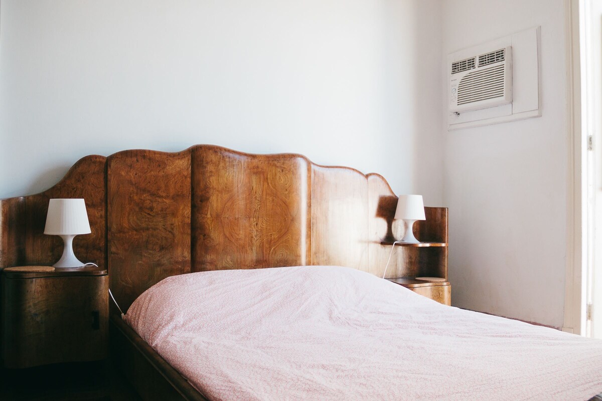 A bedroom with a wooden headboard, a pink bedspread, two bedside lamps, and a wall-mounted air vent.