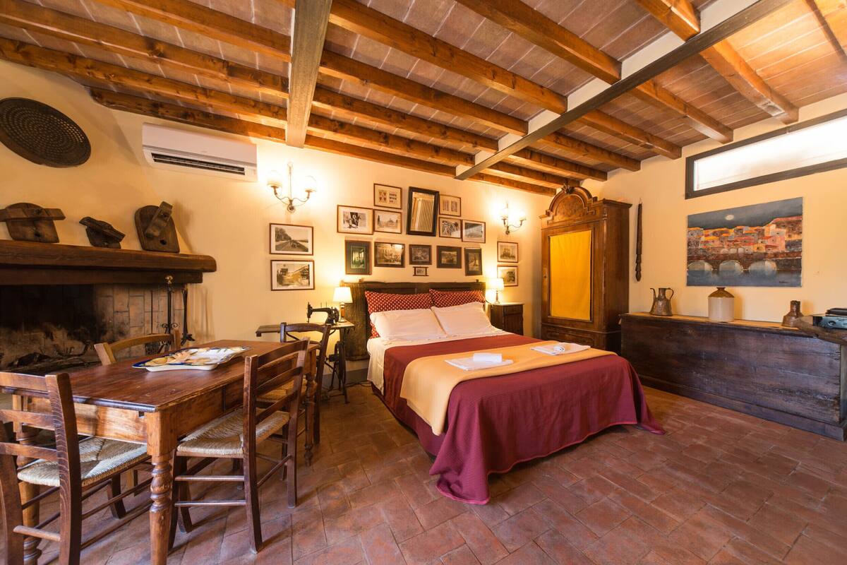 A rustic room with a wooden ceiling, a bed with a red bedspread, a dining table with chairs, and a fireplace.