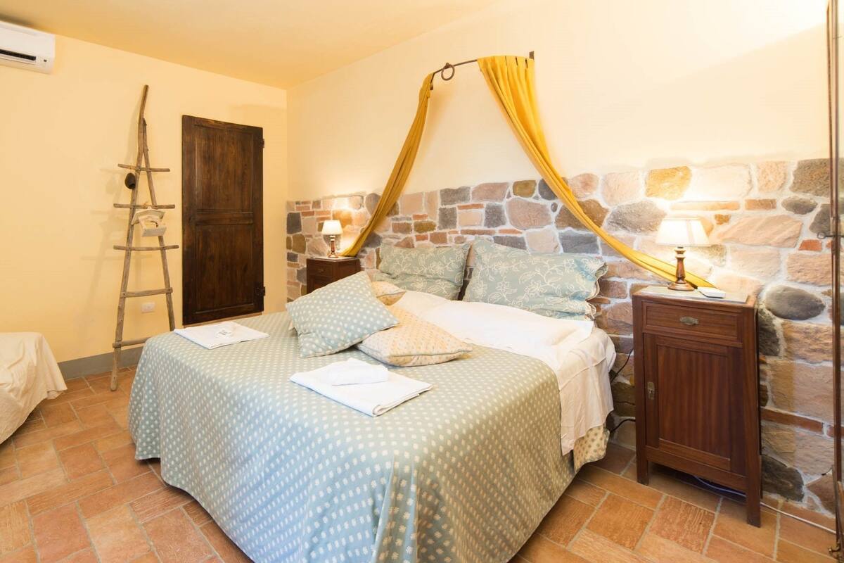 A bedroom with a bed, a wooden nightstand, a stone wall, a wooden door, and a ladder.