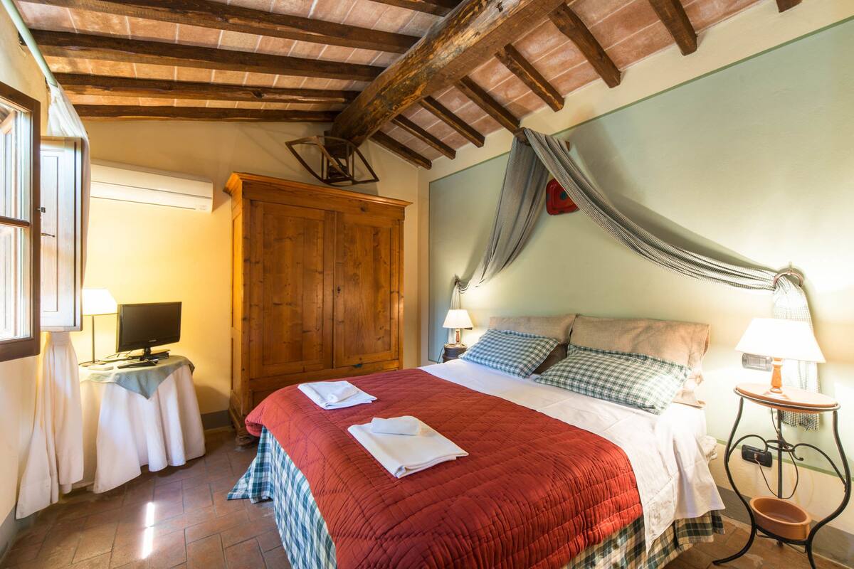 A bedroom with a wooden ceiling, a bed with a red quilt, two pillows, a nightstand with a lamp, and a wooden wardrobe.
