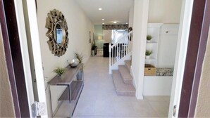 A hallway with a staircase leading to a landing. There is a decorative mirror on the wall, a console table with a vase and plants, and a white staircase with a handrail. - Aco234120 - Windsor At Westside Resort - 4 Bed 3 Baths Townhome (Kissimmee)