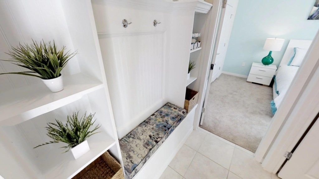 A narrow hallway with white walls and a tiled floor, leading to a bedroom with a bed, nightstand, and a green lamp.