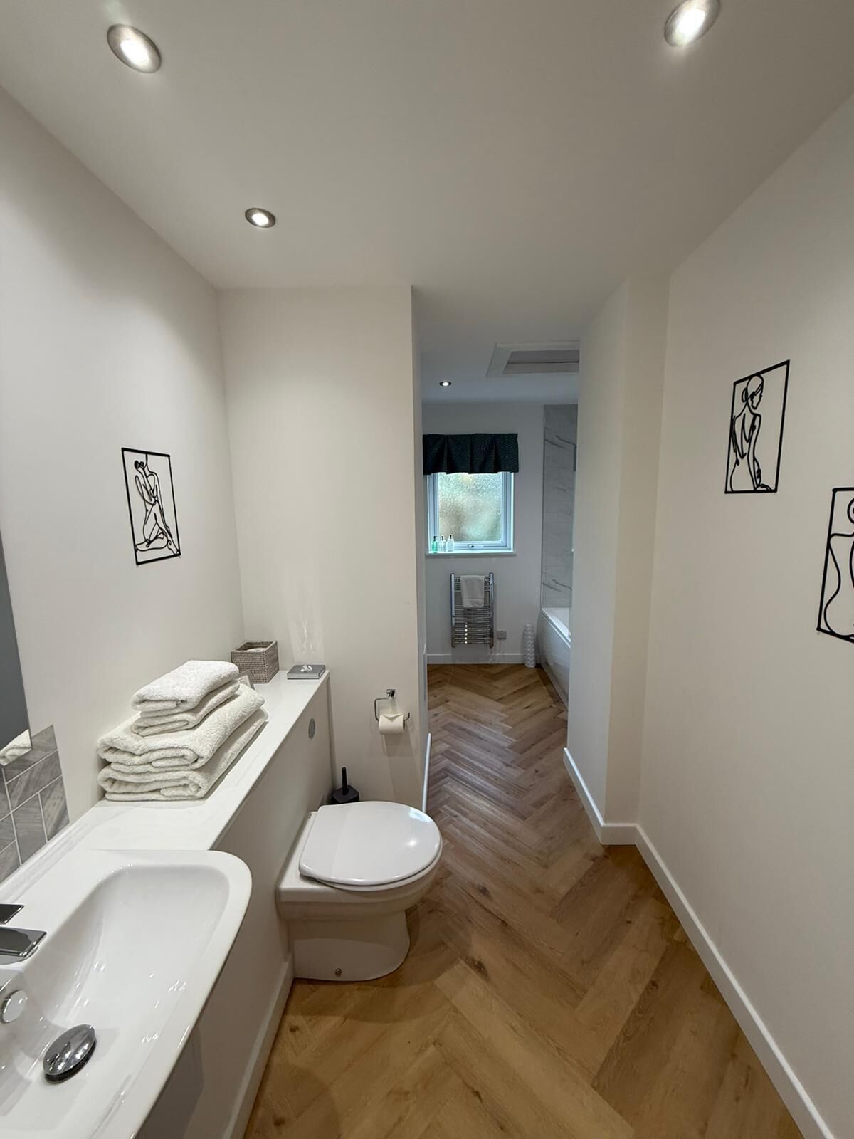 Un baño moderno con lavamanos, inodoro y suelo de madera en patrón de espiga. Hay obras de arte enmarcadas en las paredes y una ventana con vistas al exterior.