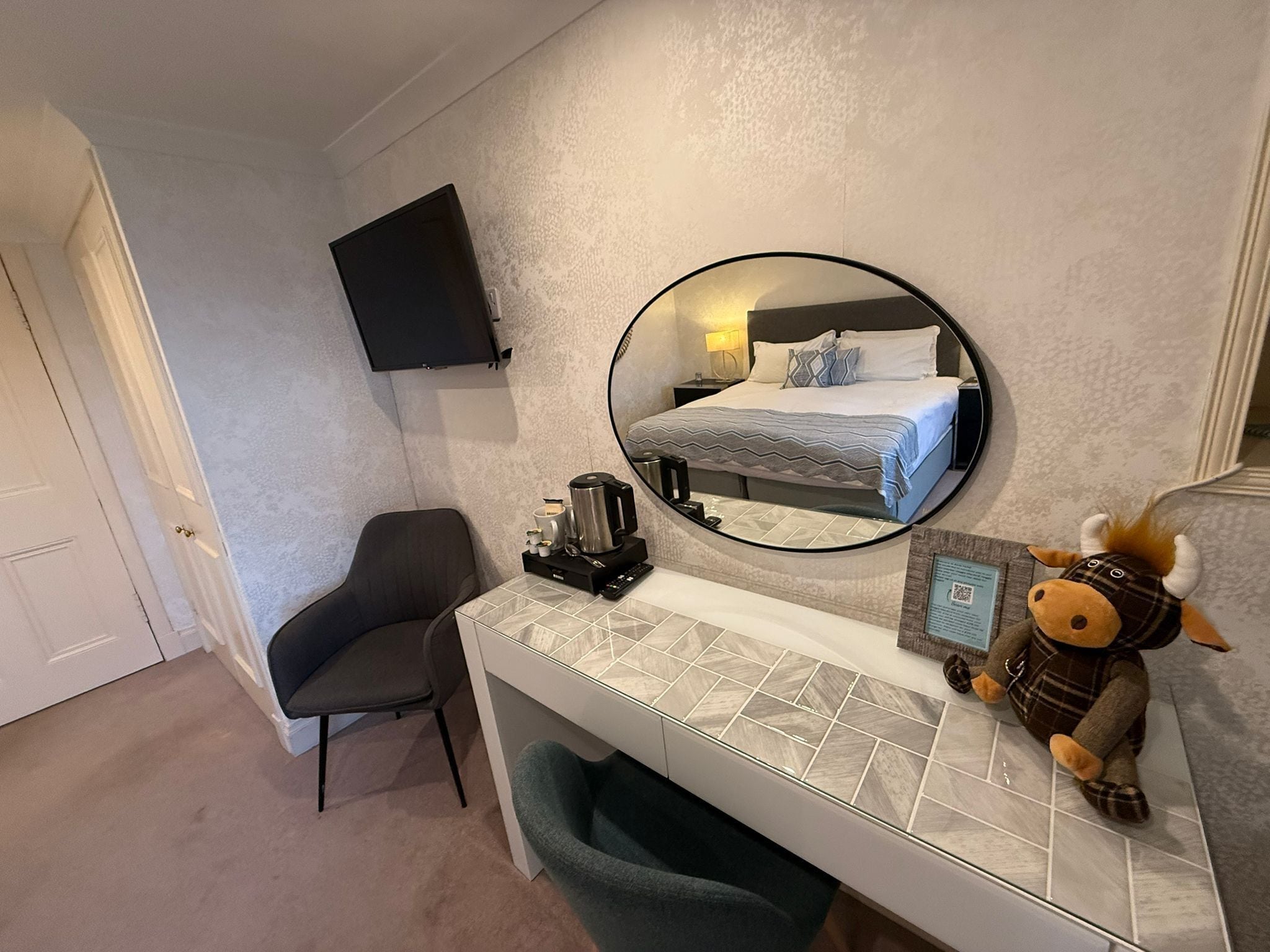 Habitación de hotel con cama, escritorio con cafetera, silla, televisor montado y un peluche en la cama.