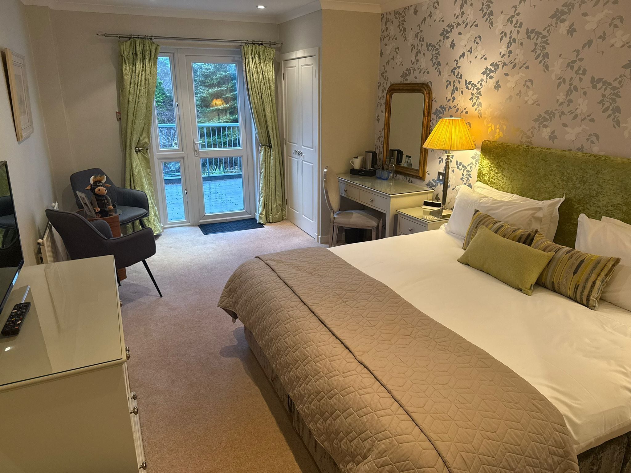 Quarto de hotel com uma cama grande, uma escrivaninha, uma cadeira e vista para o exterior através de uma porta de vidro.