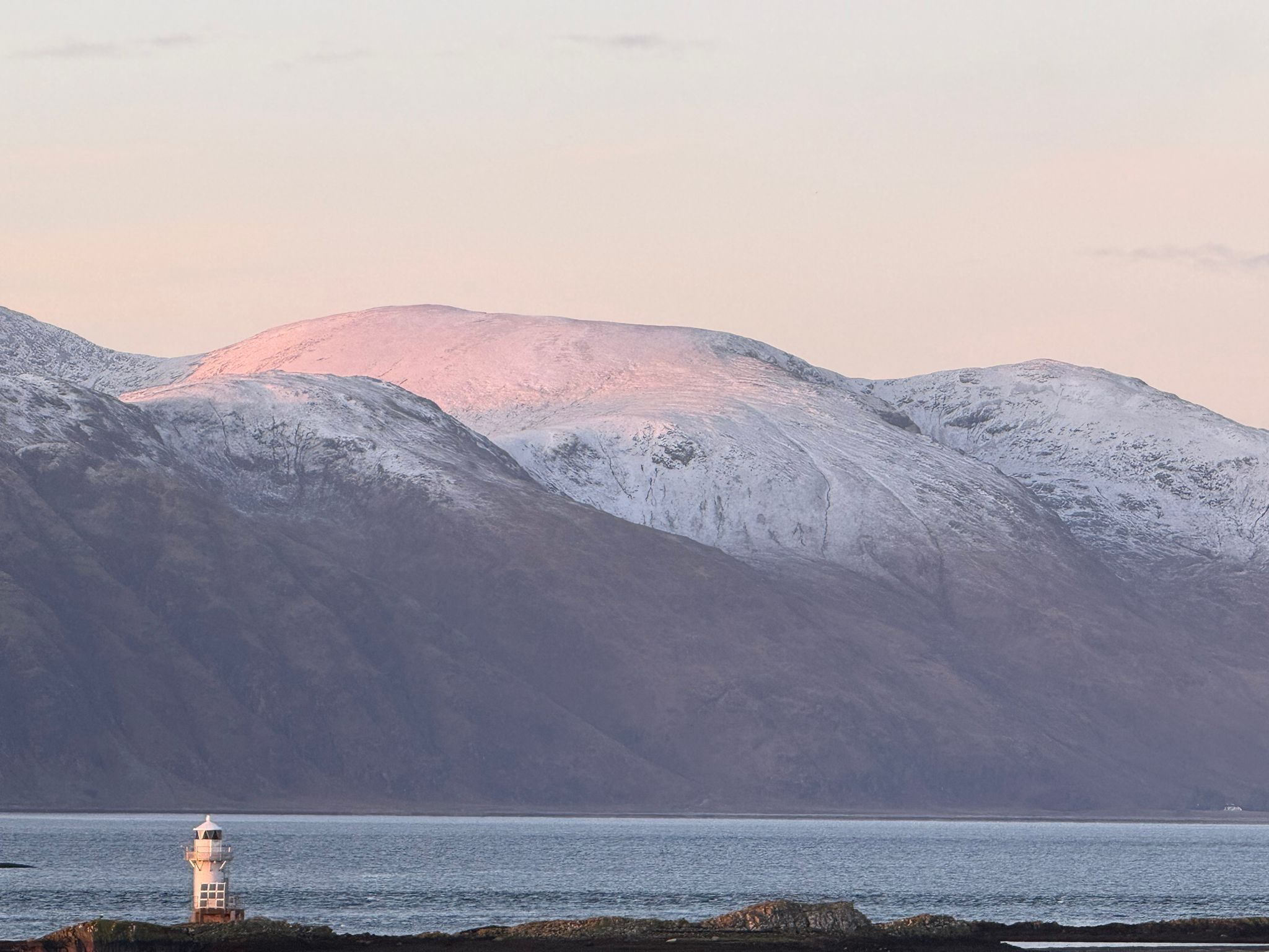 Un faro junto a un mar tranquilo, con montañas nevadas al fondo.