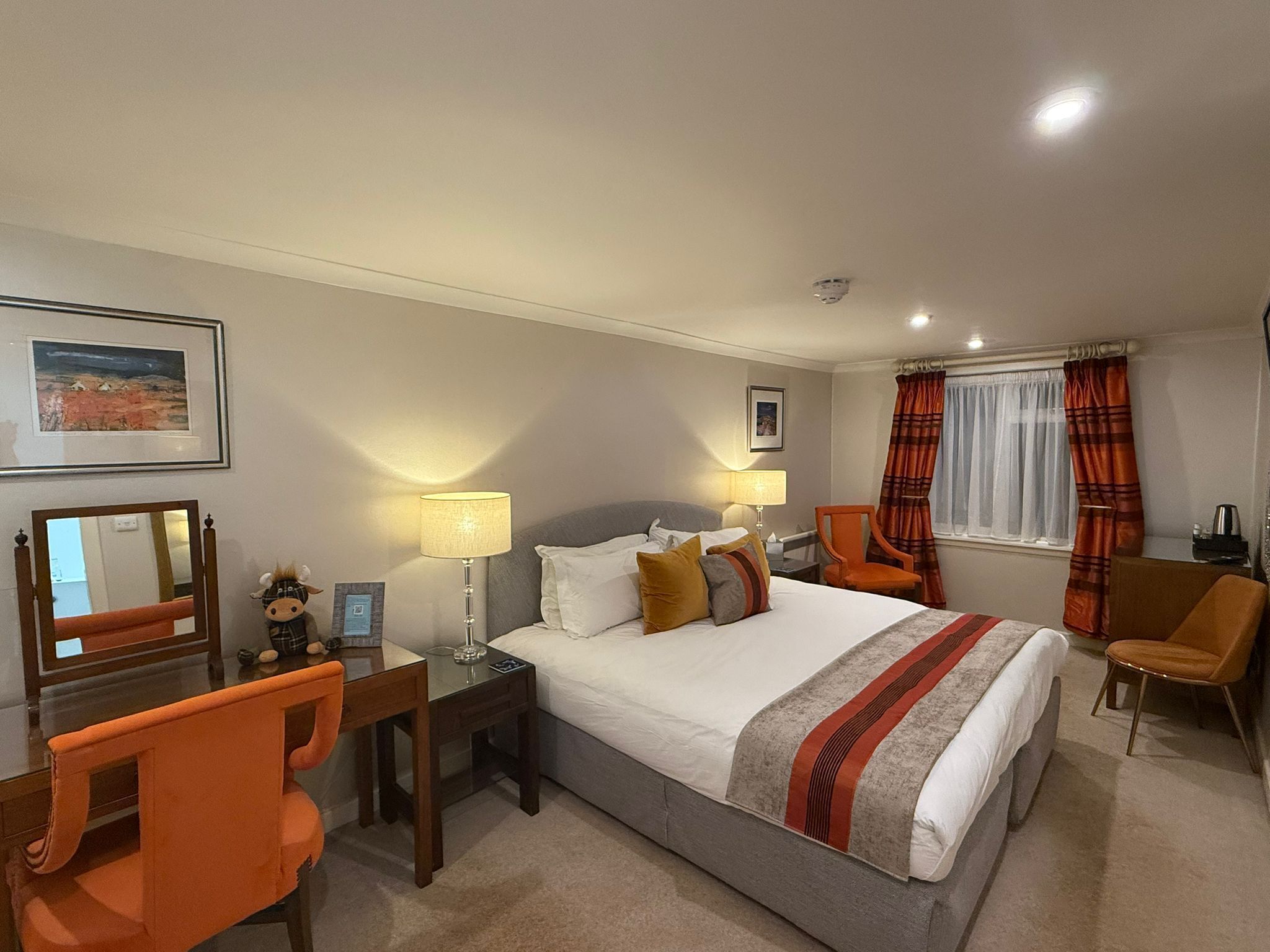A hotel room with a large bed, two bedside tables with lamps, a desk with a chair, and a window with curtains.