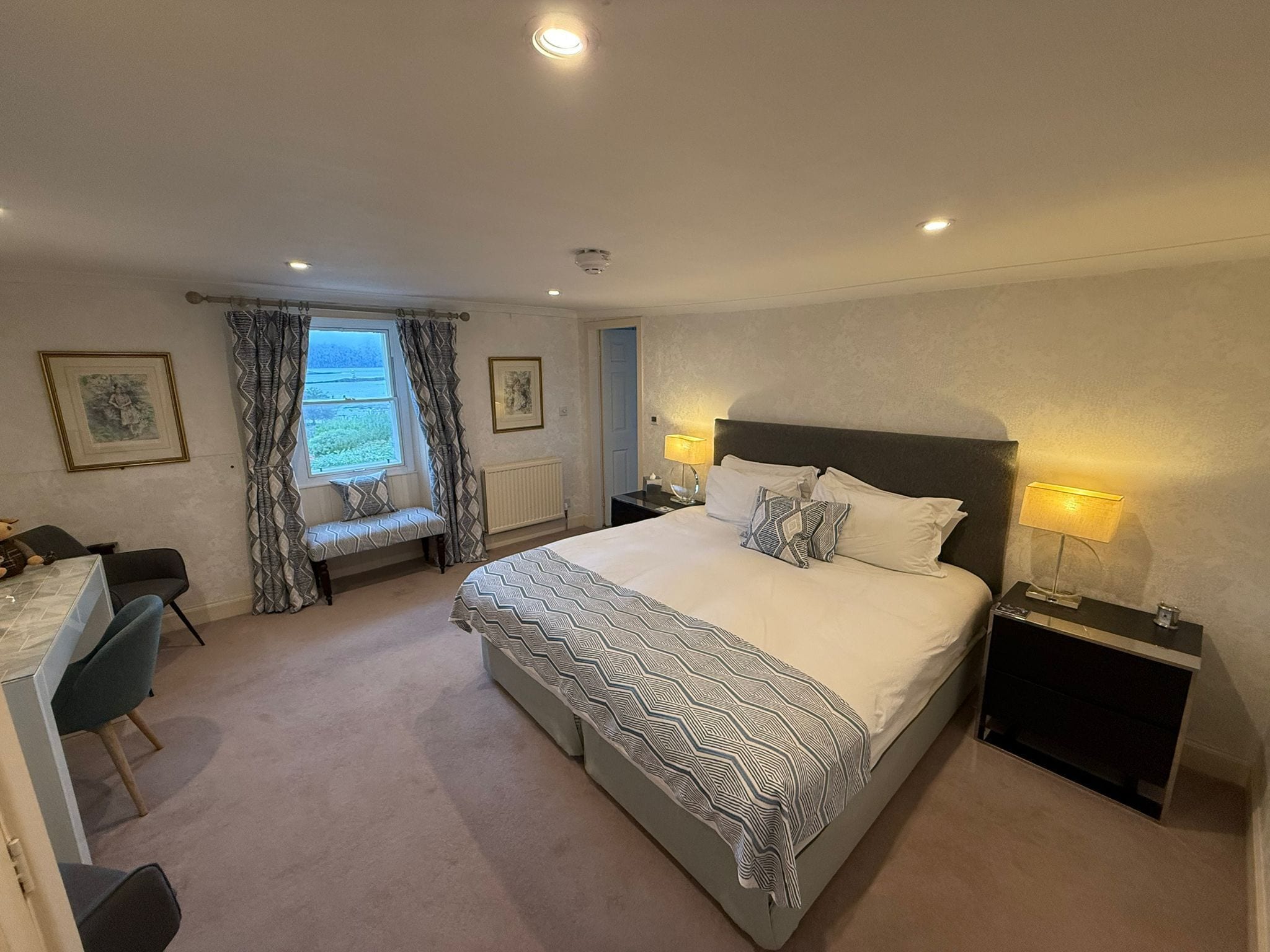 A bedroom with a bed, bedside tables, a bench, a desk, and a window with curtains.