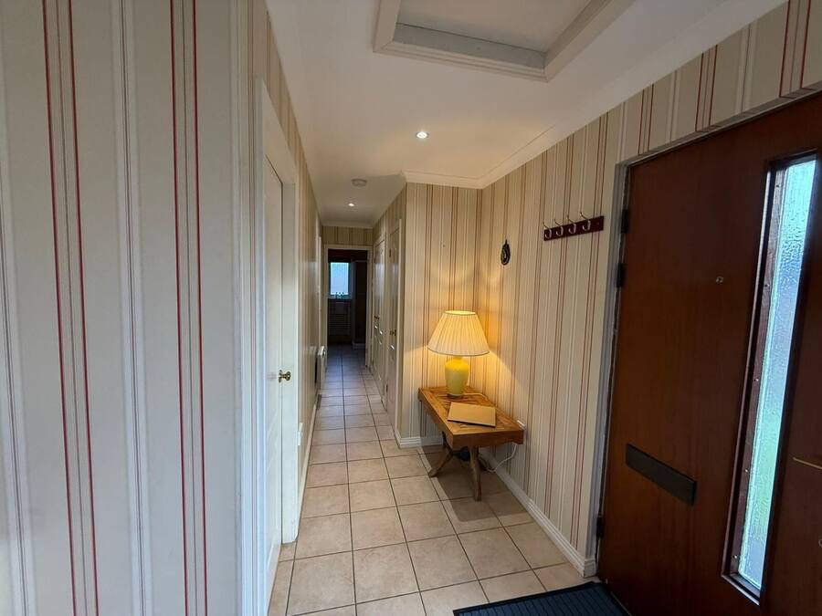 Un pasillo con papel tapiz a rayas, una puerta de madera, una mesita con una lámpara y una ventana.