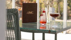 A glass table with a menu, two drinks, and a view of trees through large windows. - The Airds Hotel & Restaurant (Appin)