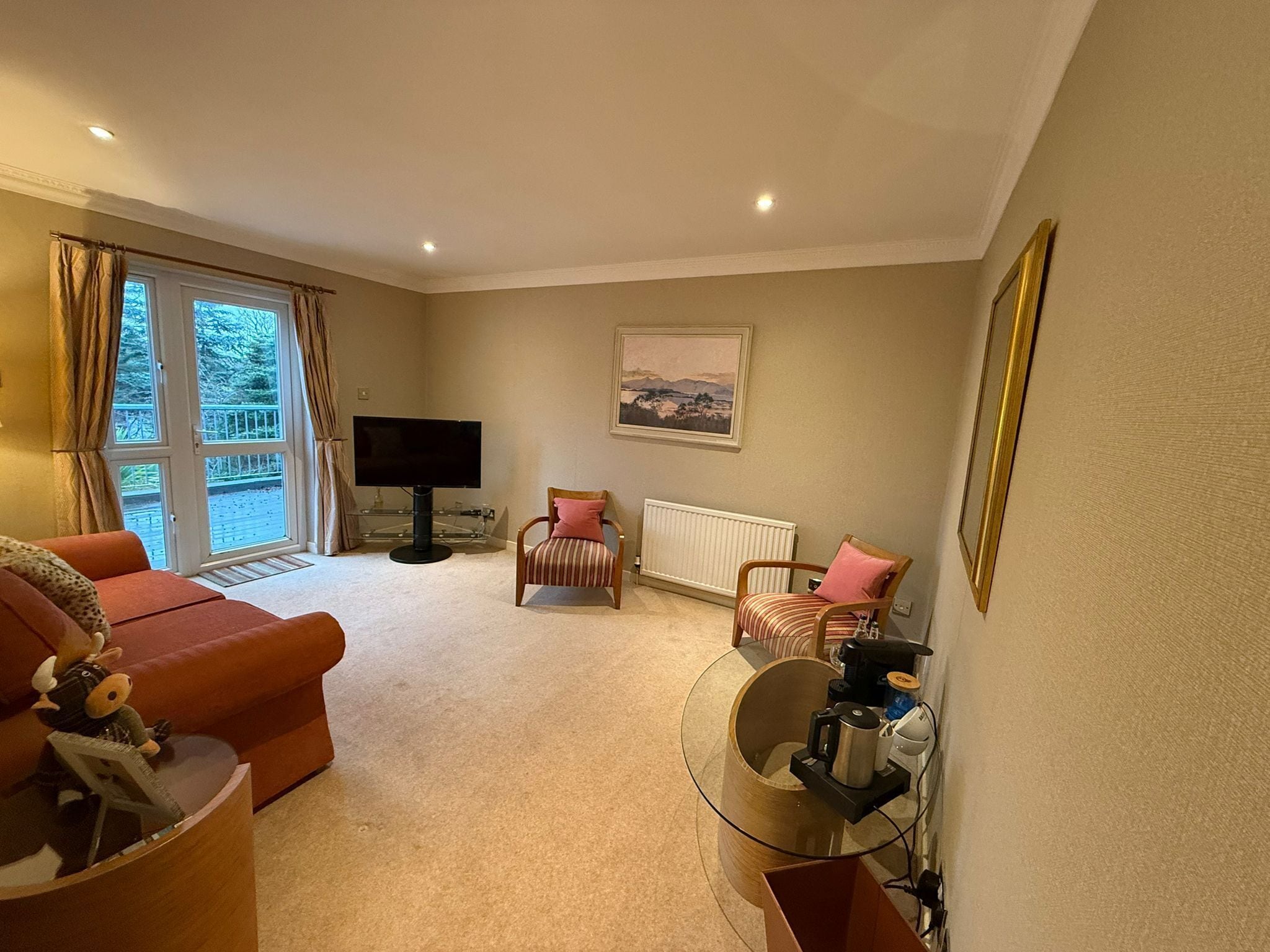 A living room with a sofa, armchair, and a television. There is a window with curtains, a radiator, and a painting on the wall.