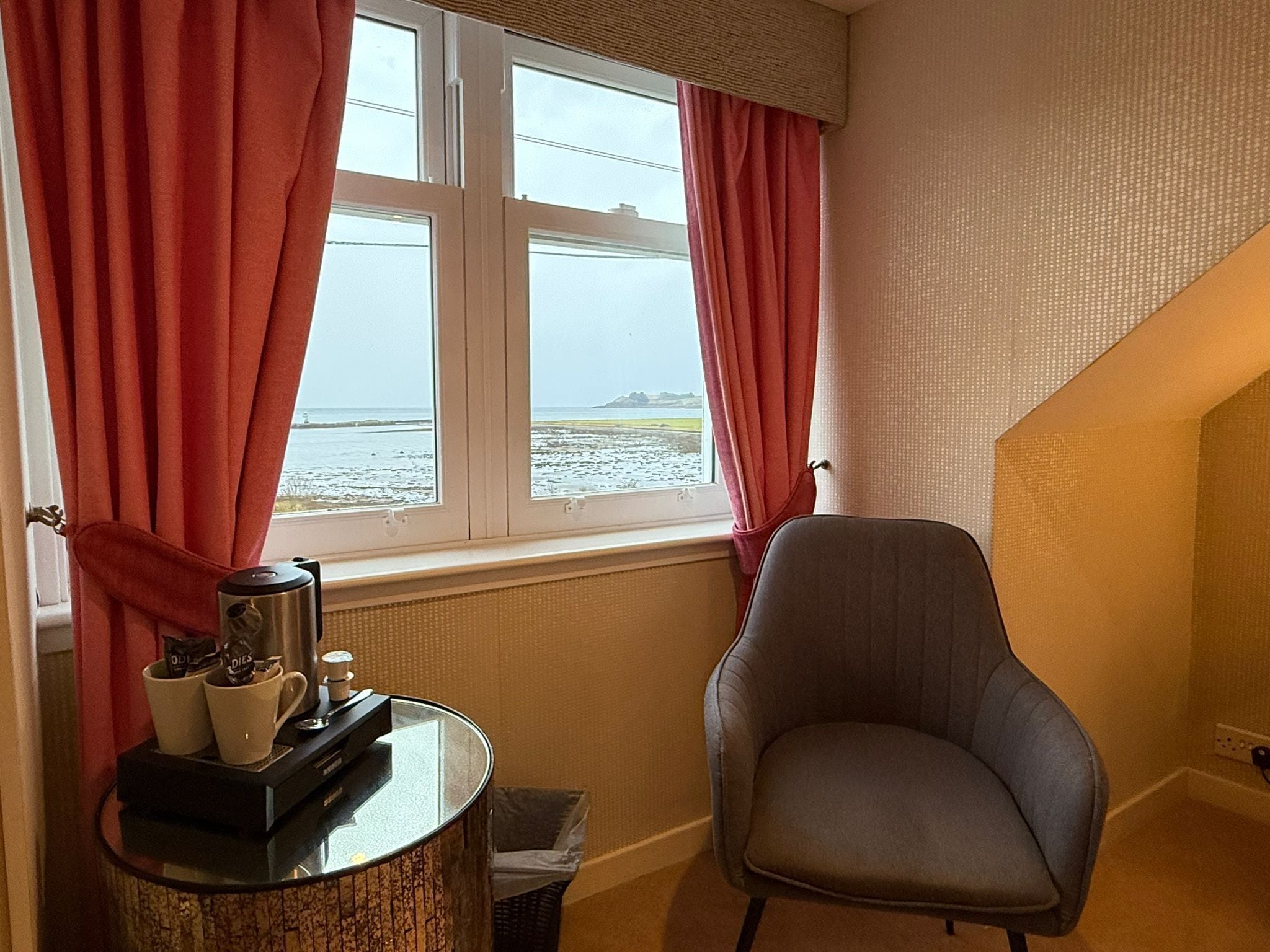A room with a view of the sea, a chair, a table with a coffee maker, and a radiator.