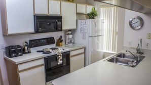 Una cocina compacta con gabinetes blancos, un microondas negro, un refrigerador de acero inoxidable, un horno empotrado, una cafetera y un lavamanos con grifo.