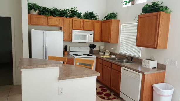 A compact kitchen with wooden cabinets, a white refrigerator, a microwave, a stove, a sink, and a dishwasher. There are potted plants on the cabinets and a window with blinds. - Ahr225 - Southern Dunes - 3 Bed 2 Baths Villa (Haines City)