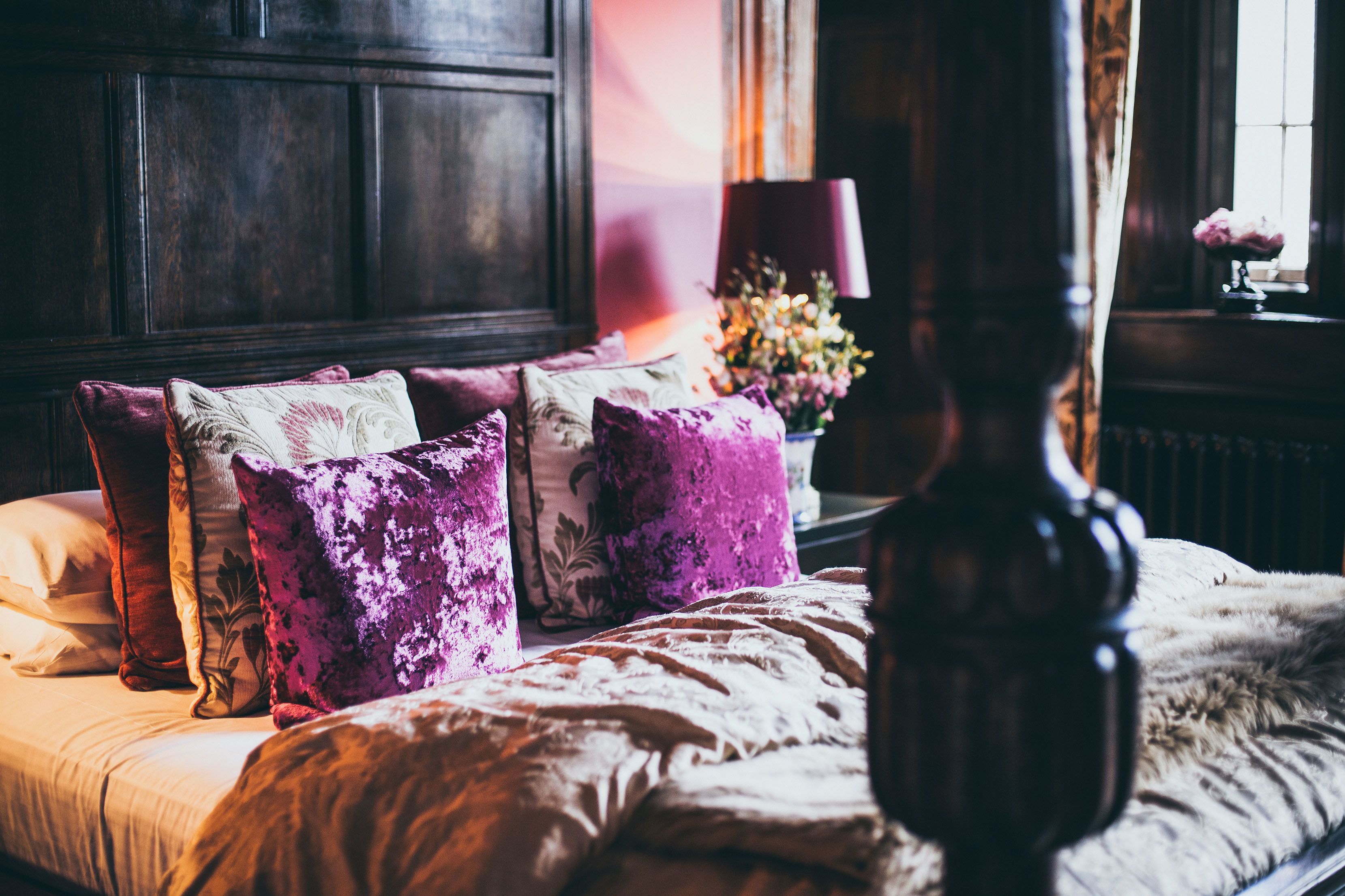 A bed with multiple pillows, a wooden headboard, and a nightstand with a lamp and flowers.