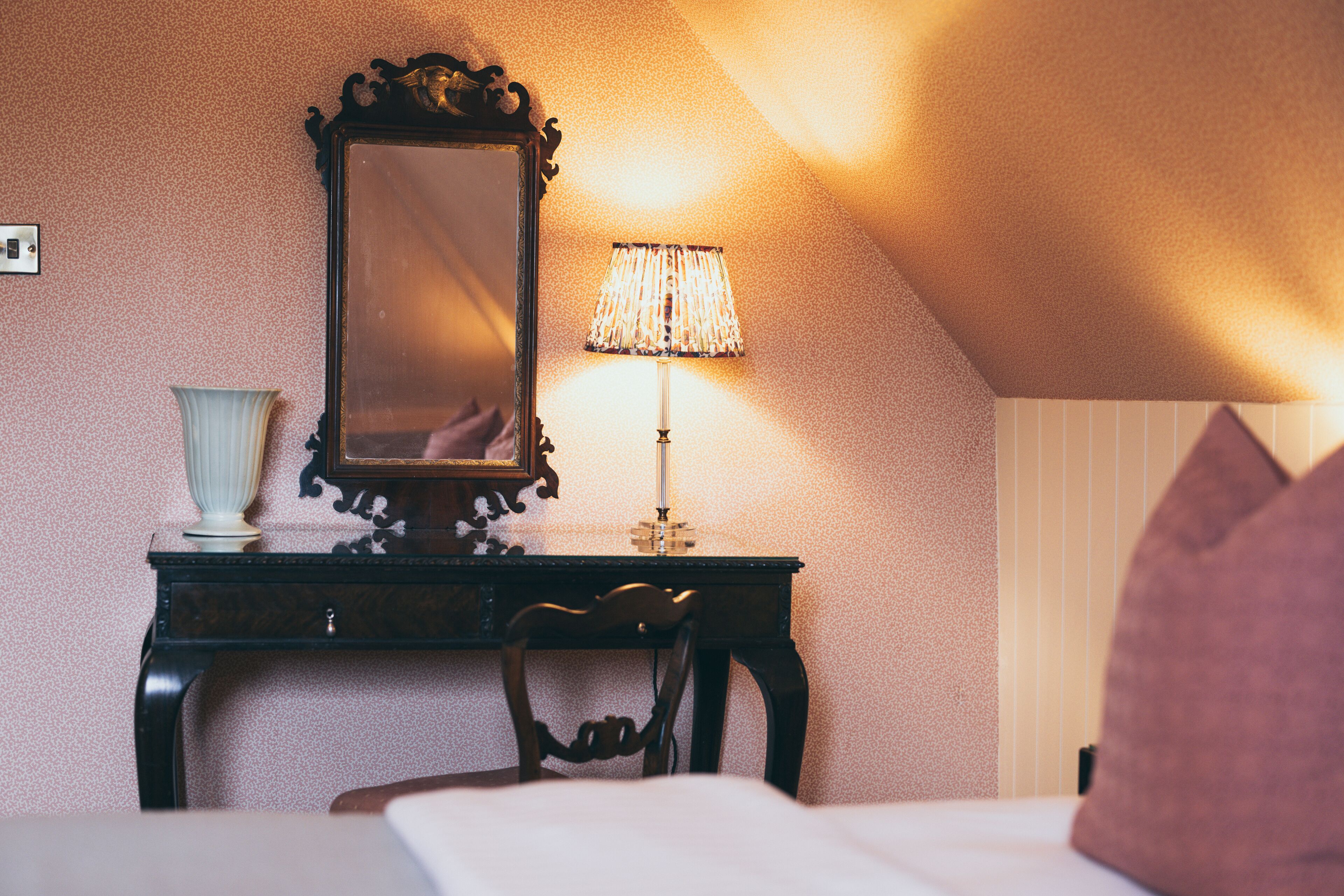 A bedroom with a bed, a wooden nightstand, a lamp, a mirror, and a vase.
