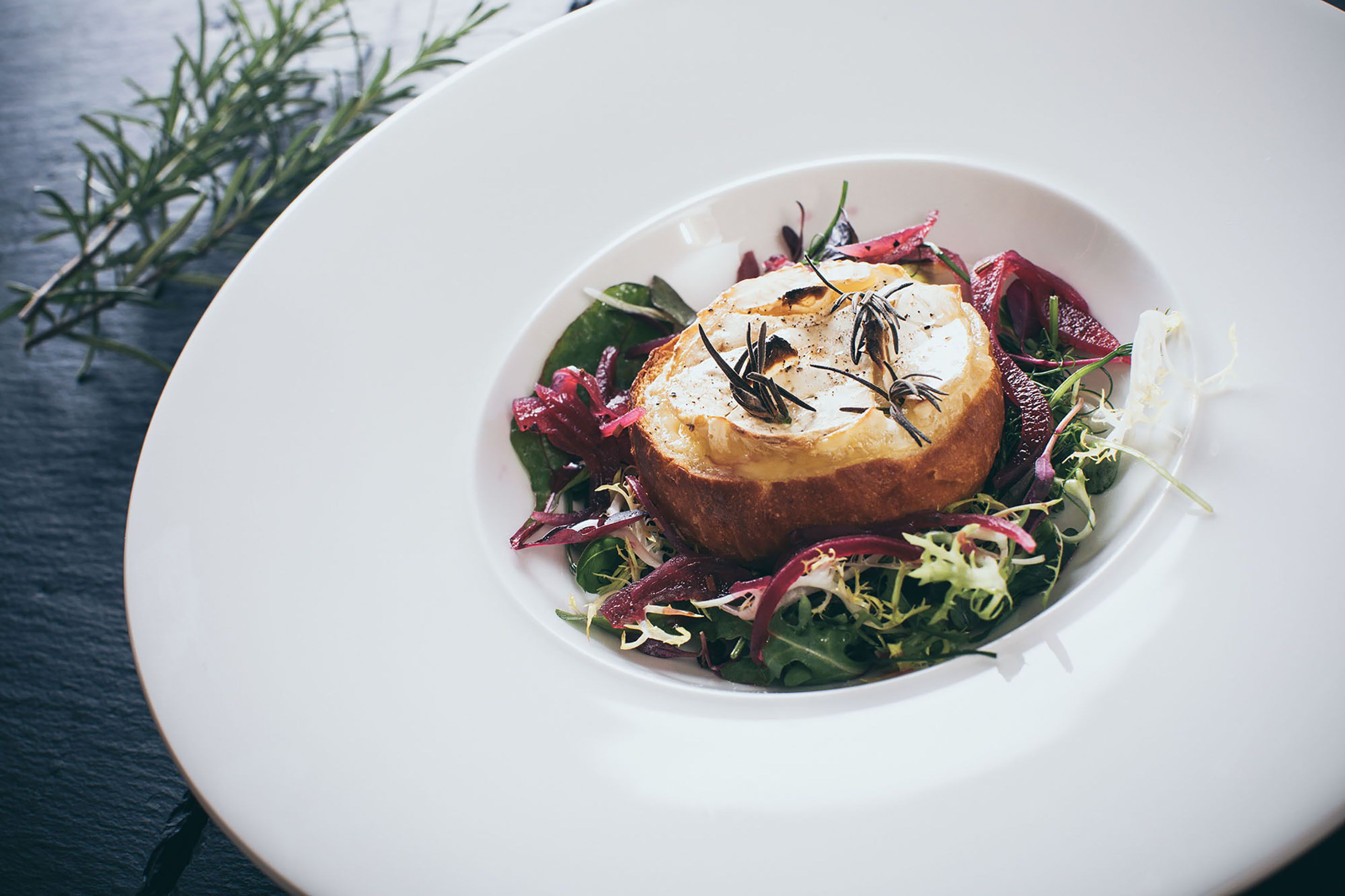 A white plate with a round, toasted bread topped with herbs, surrounded by mixed greens and red leaves.