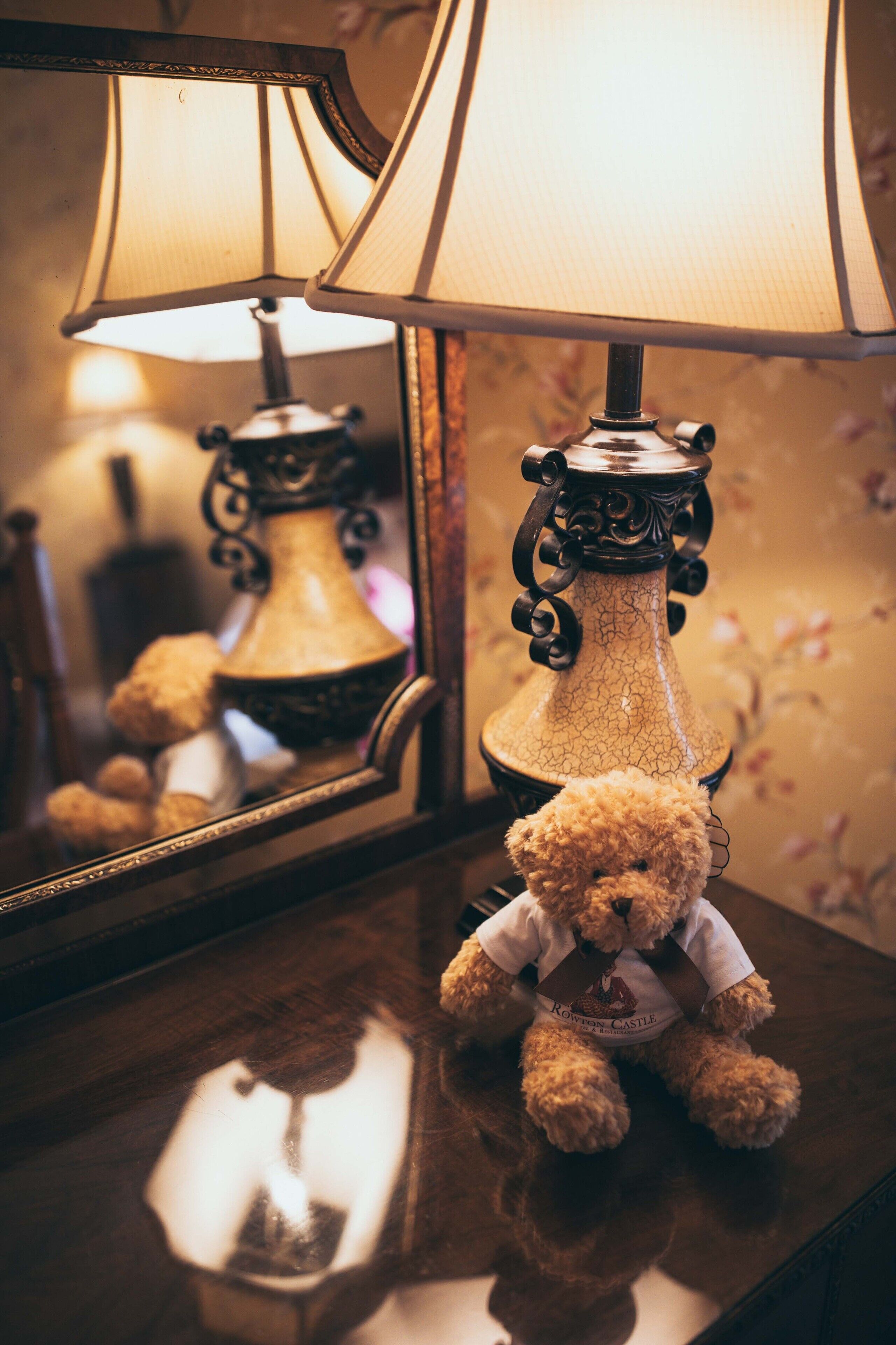 A teddy bear on a wooden surface with a lamp and mirror behind it.
