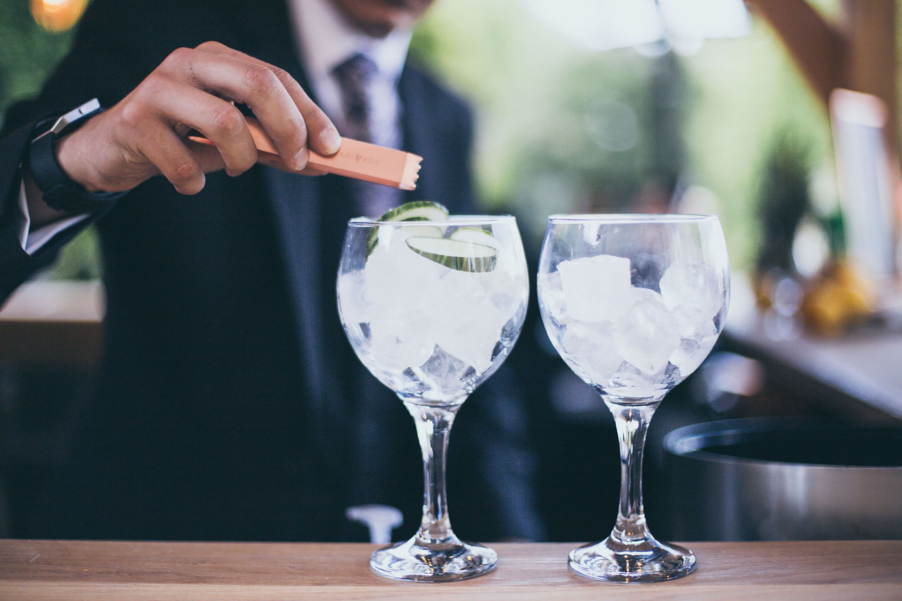 Una persona vestida con traje sirve una bebida en un vaso con hielo y una rodaja de pepino.