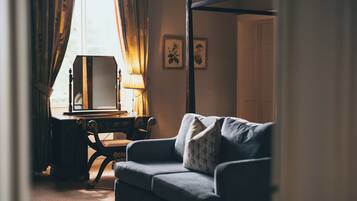 A room with a four-poster bed, a sofa, a desk with a lamp, and two framed pictures on the wall.