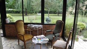 A covered patio with two chairs, a table with magazines, and a view of a garden through large windows. - Chambres d'hôtes La Planche Pinçon (Teloché)