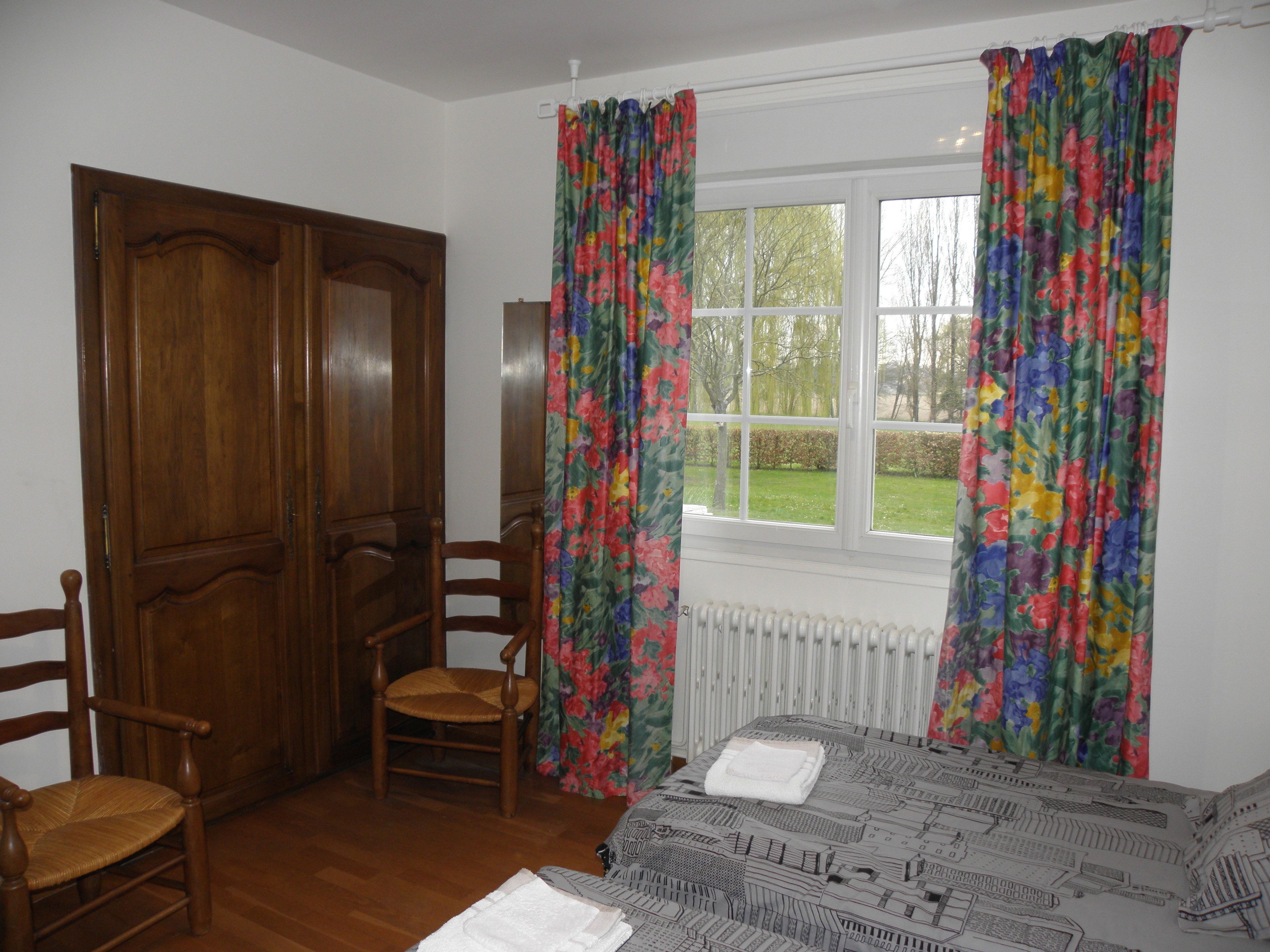 A bedroom with a wooden wardrobe, a chair, a bed with a patterned bedspread, and a window with floral curtains.