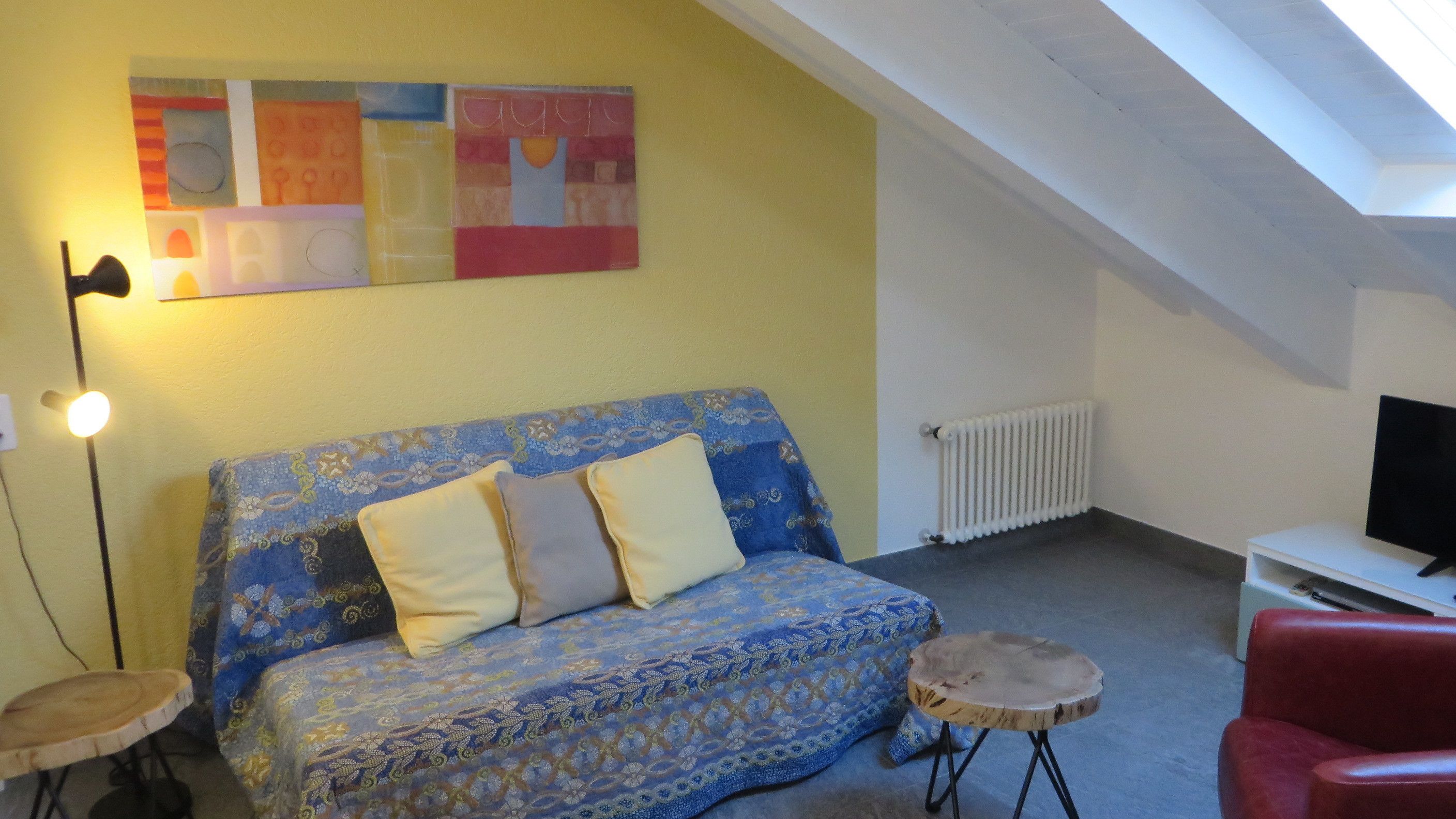 Une pièce chaleureuse avec un canapé à motifs, une petite table ronde, un fauteuil rouge, un téléviseur et un radiateur.