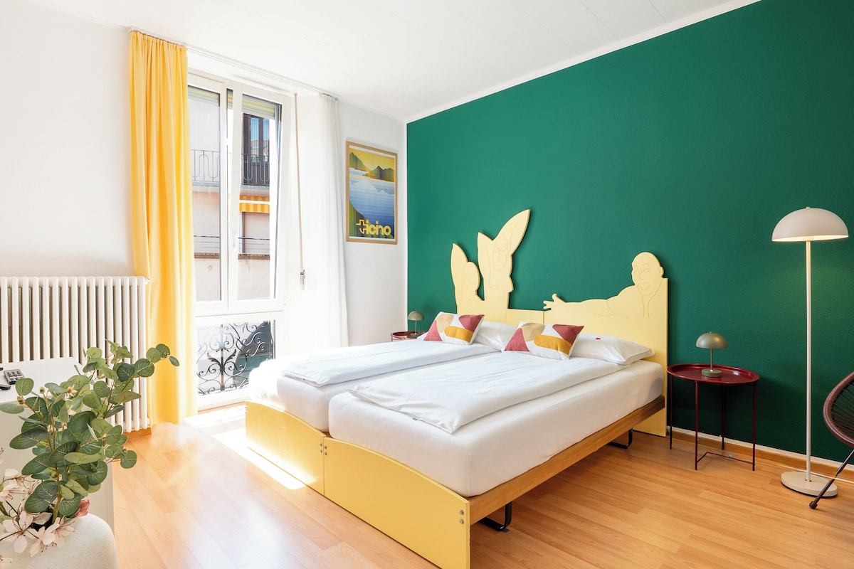 A hotel room with a bed, a nightstand, a lamp, a window with curtains, and a green wall.