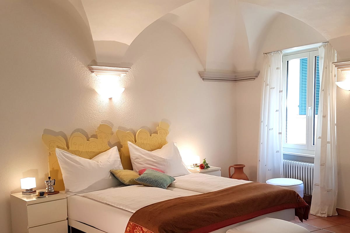 Un dormitorio con cama, lámparas de noche, mesita de noche, ventana con cortinas y un radiador.