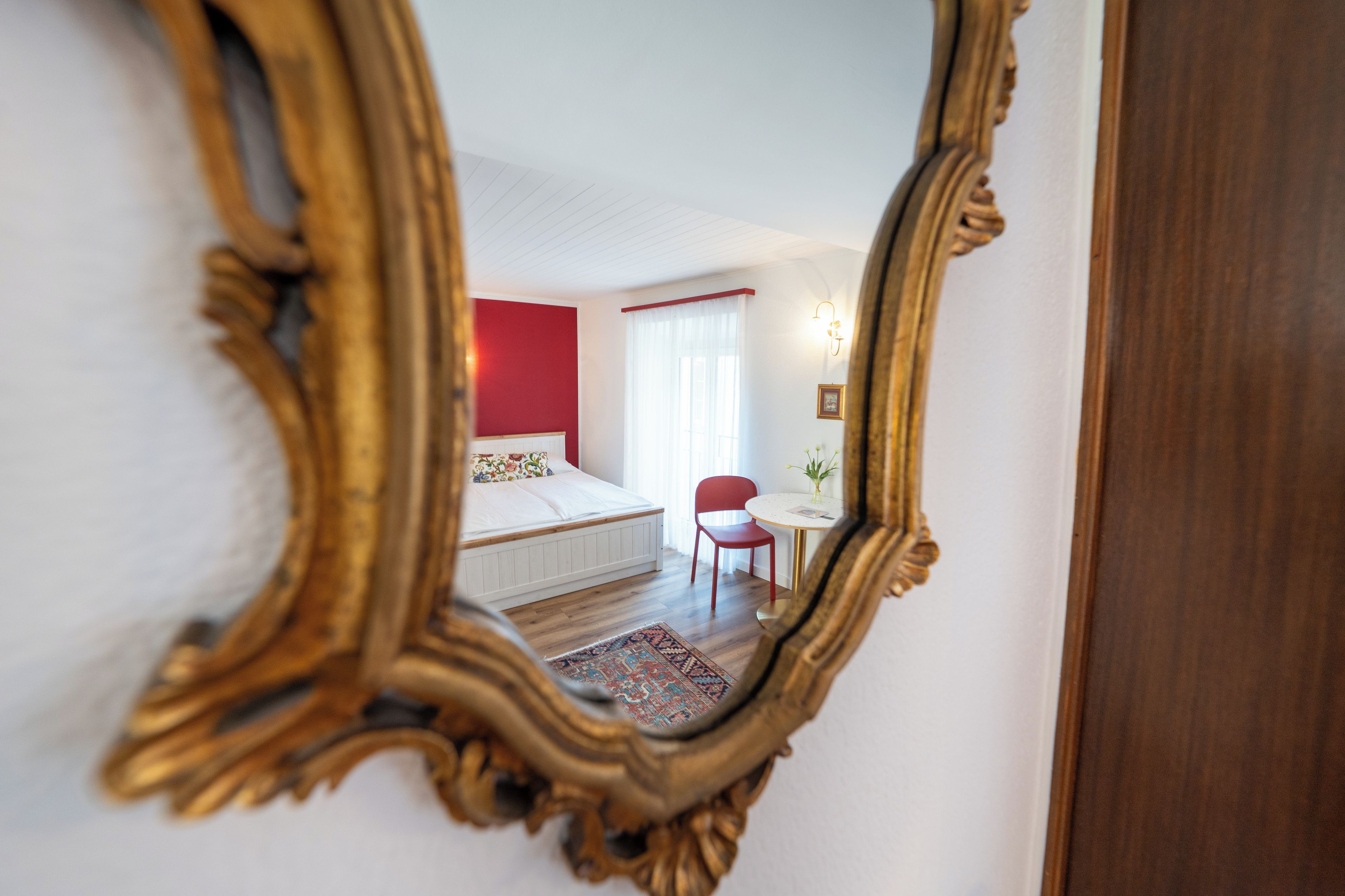 A bedroom with a bed, a chair, a small table, and a mirror reflecting the room.