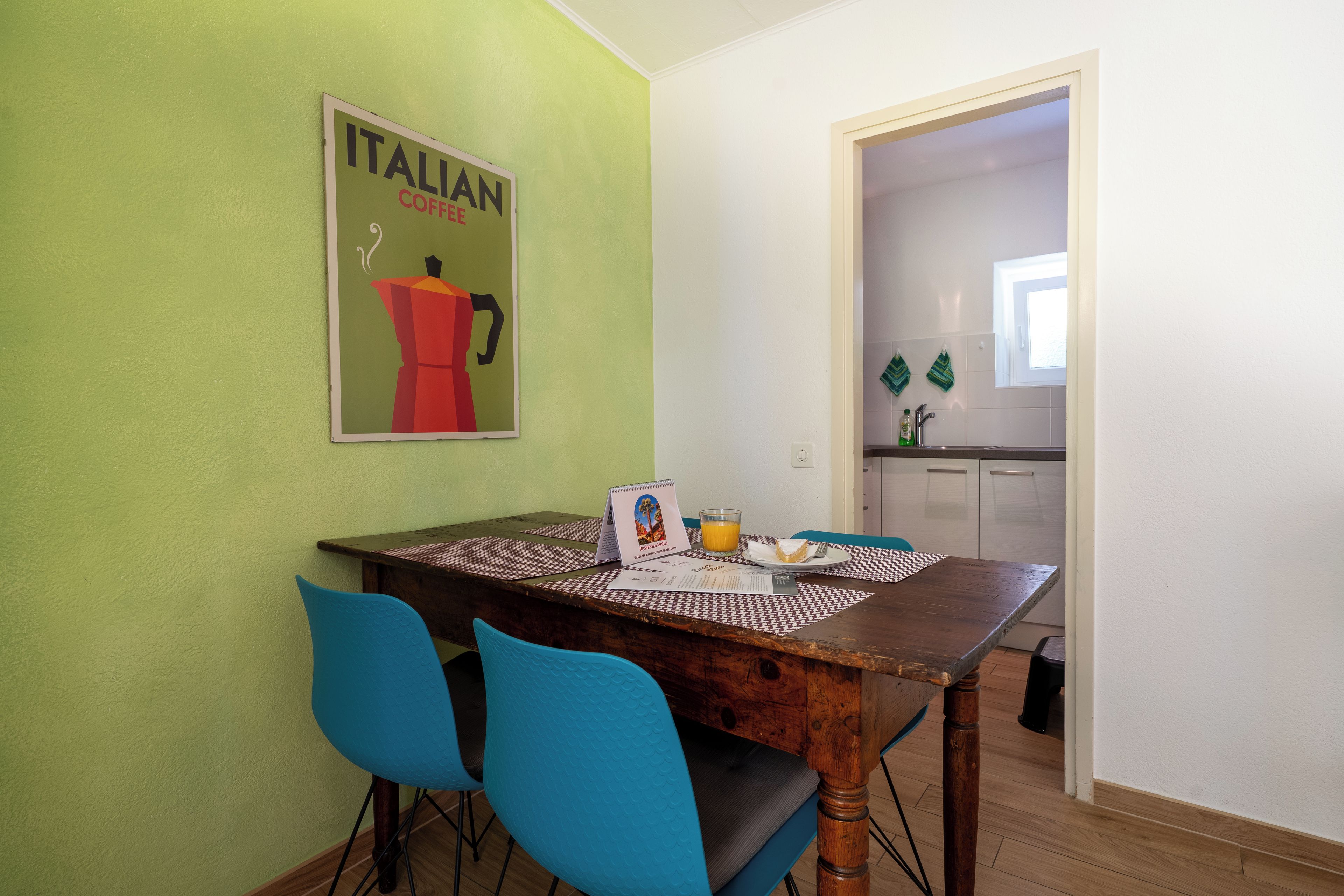 Ein Essbereich mit einem Holztisch, zwei blauen Stühlen, einem Poster an der Wand und einem Blick auf eine Küche im Hintergrund.