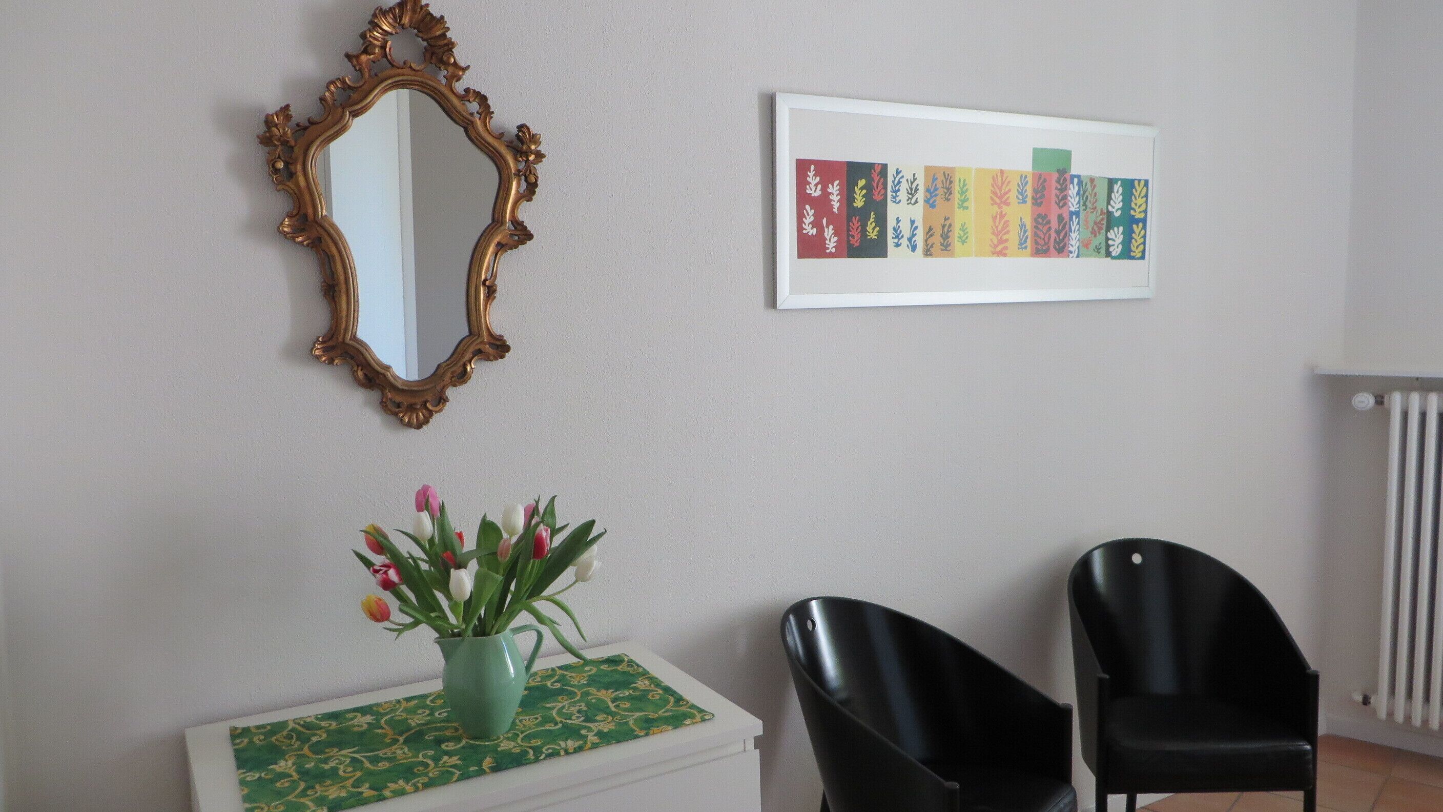 Una habitación con un espejo, una obra de arte enmarcada, un jarrón con flores y dos sillas.