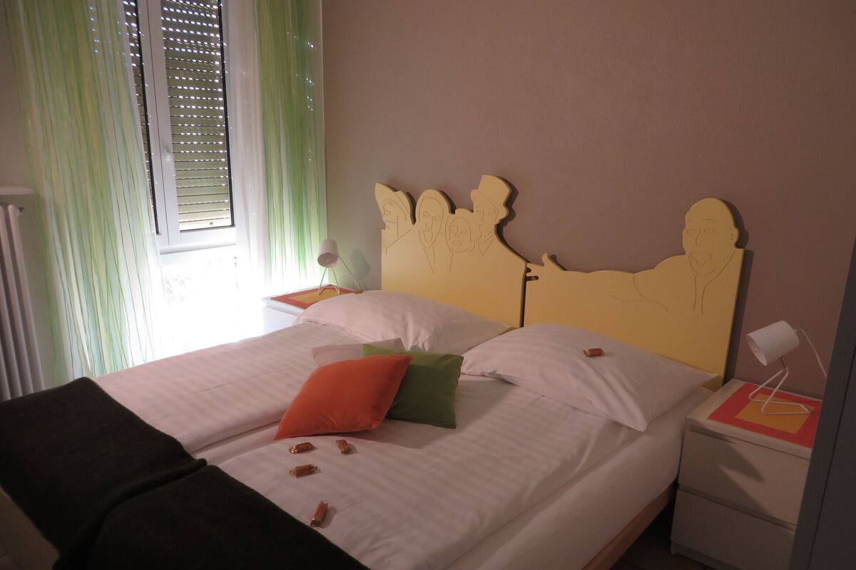 Una cama con ropa blanca, un cabecero con diseño facial y una mesita de noche con una lámpara.