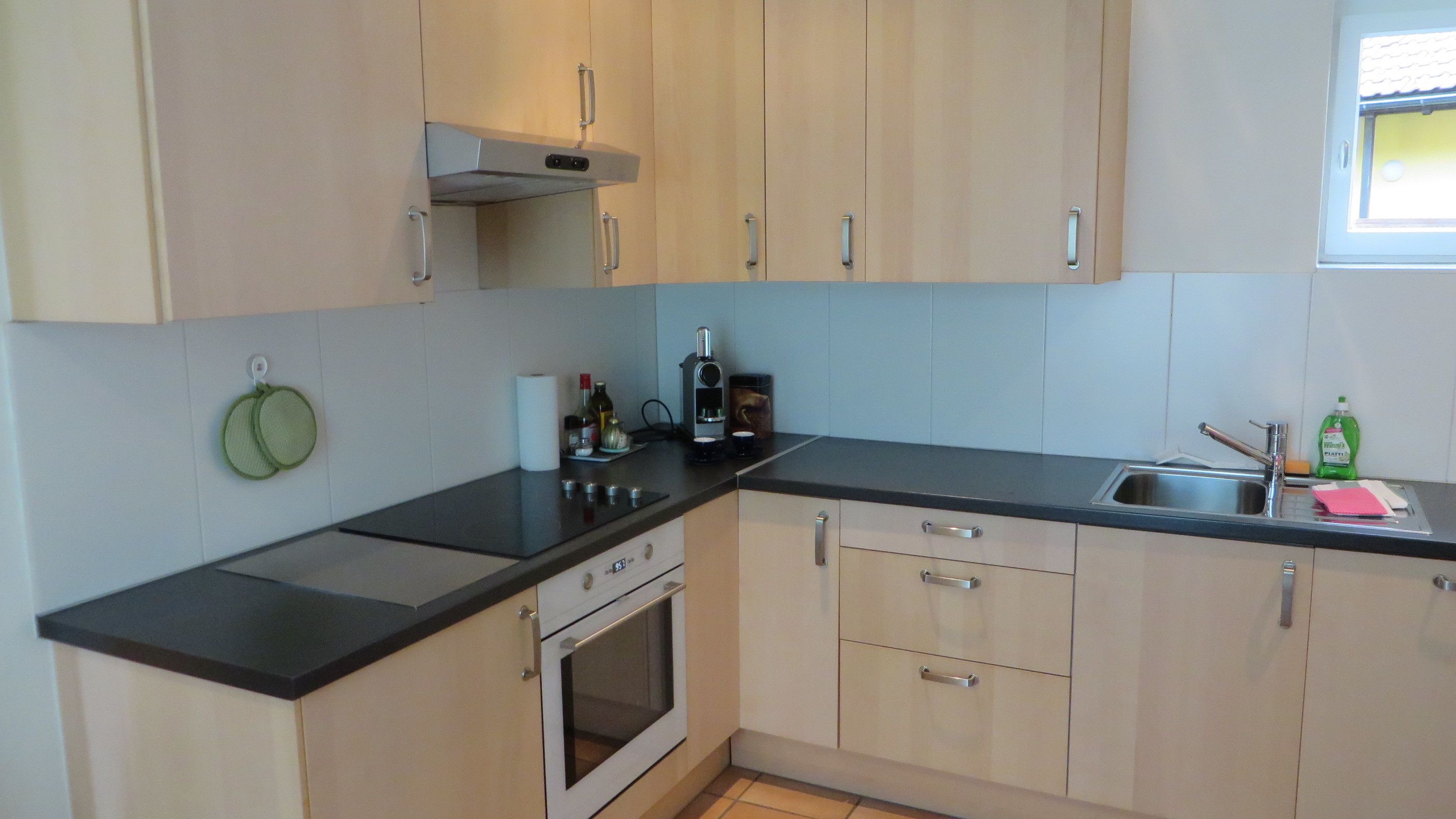 A kitchen with light wood cabinets, a black countertop, a stainless steel sink, and a built-in oven.