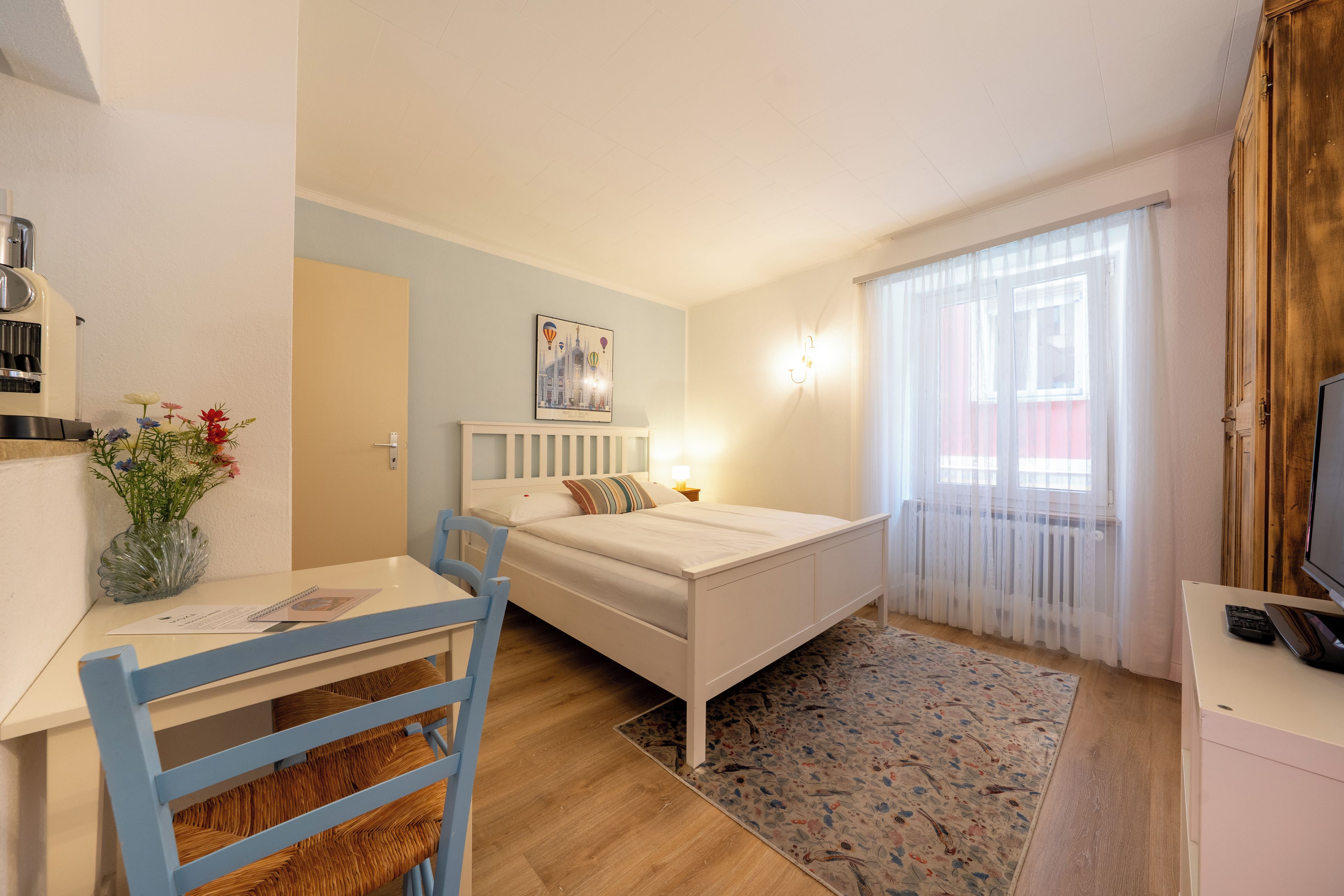 Ein gemütliches Schlafzimmer mit einem Einzelbett, einem Schreibtisch mit Stuhl, einem kleinen Tisch mit Blumenvase und einem Fenster mit leichtem Vorhang.