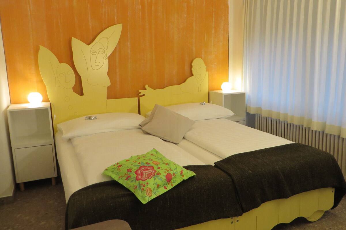 Una cama con cabecera amarilla, ropa de cama blanca y un cojín floral verde. Hay dos mesitas de noche con lámparas.