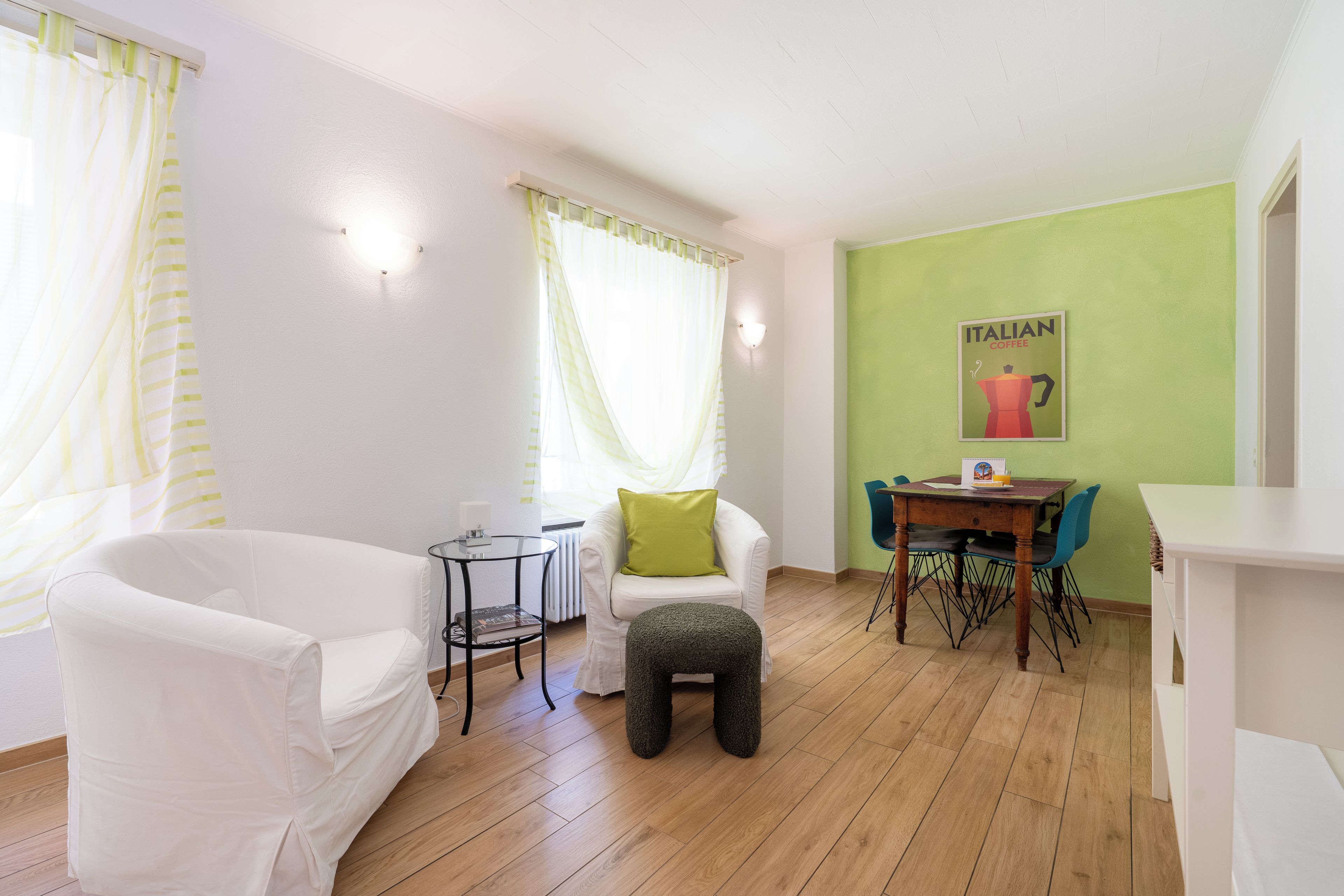 Ein Wohnzimmer mit einem weißen Sofa, einem kleinen runden Tisch, einer grünen Wand mit einem italienischen Kaffeeplakat und Holzdielen.