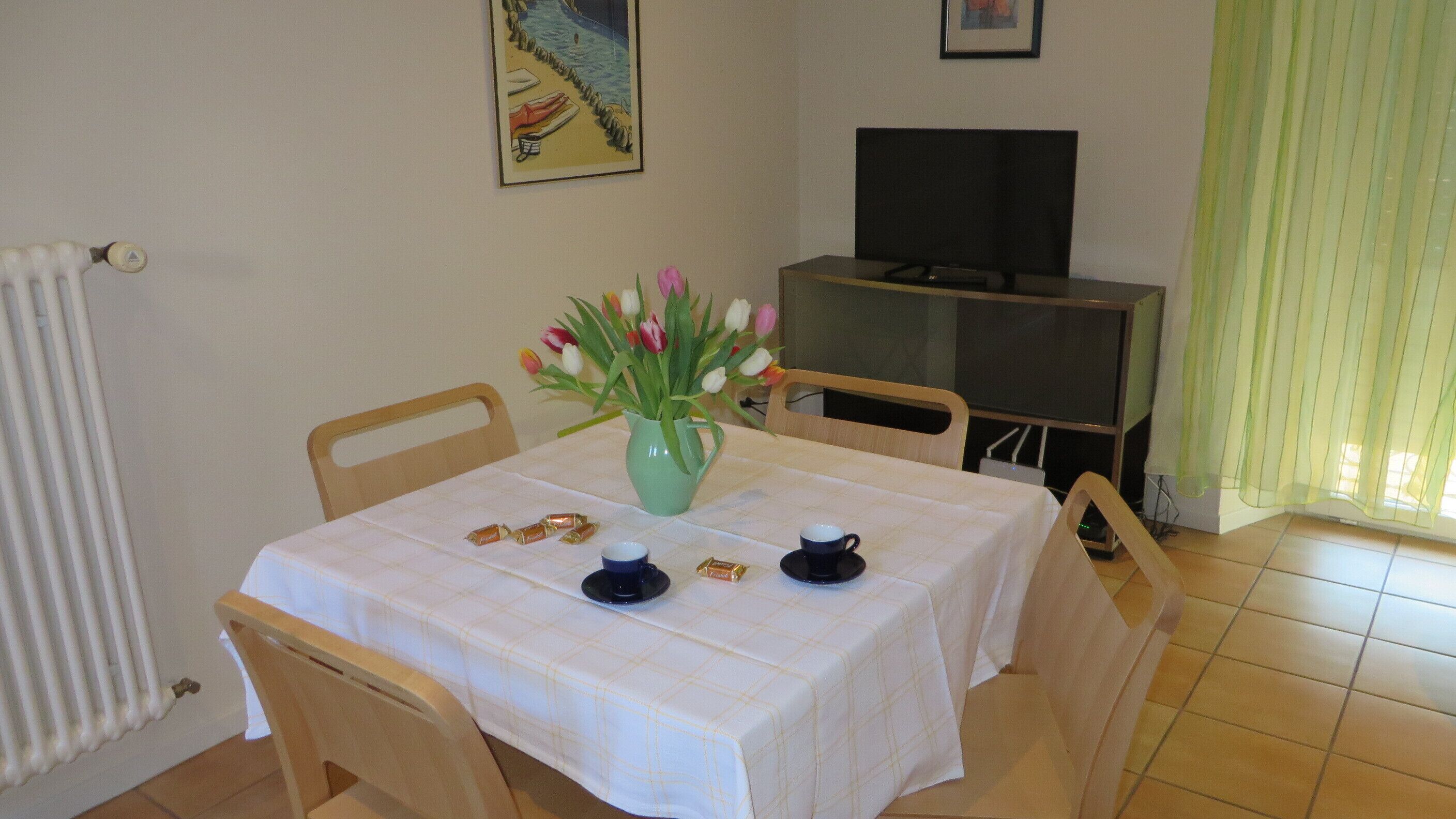 Un comedor con una mesa lista para dos personas, un jarrón con tulipanes y un televisor al fondo.