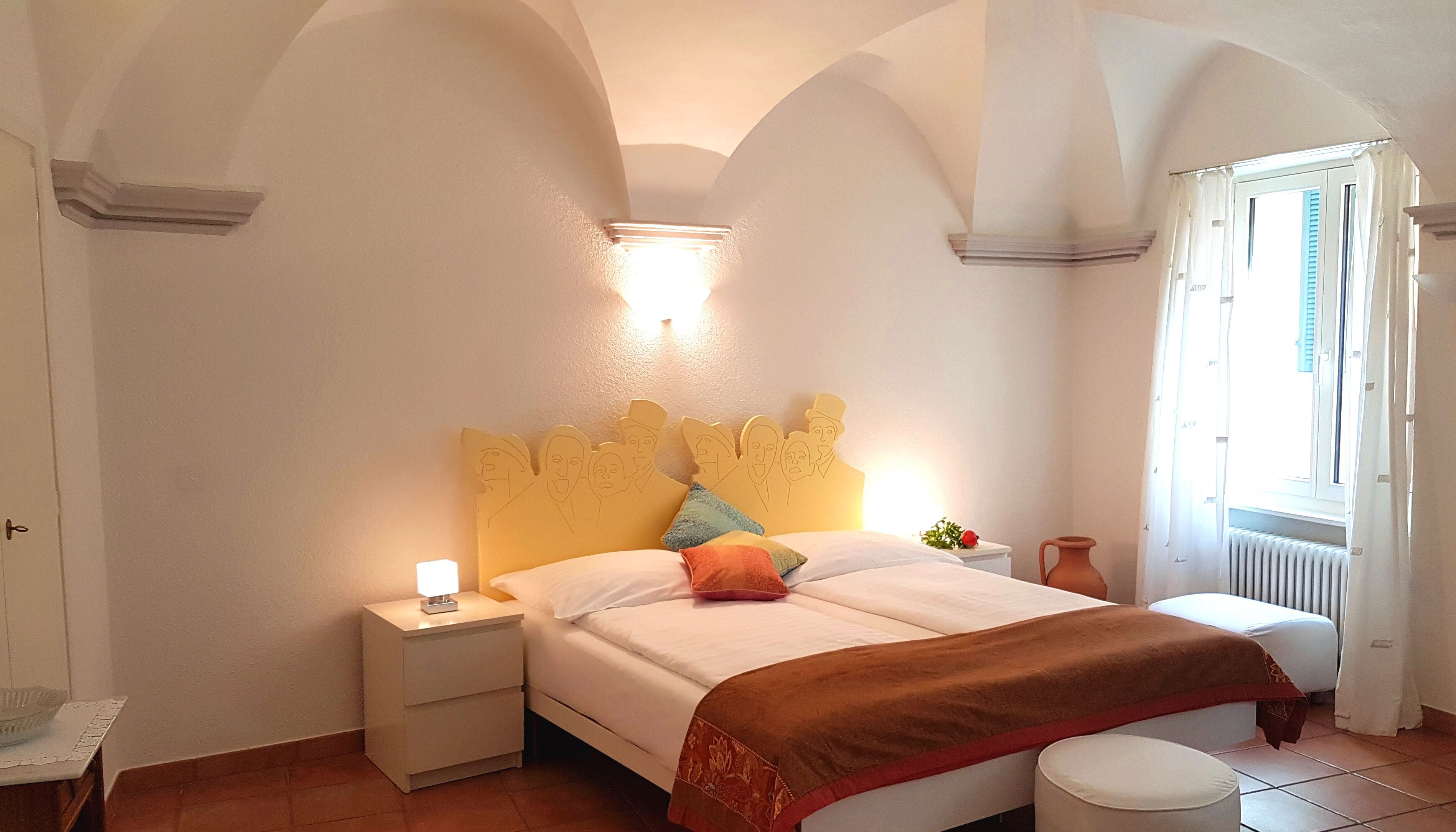 A bedroom with a bed, nightstands, a bedside table, a radiator, and a window with curtains.