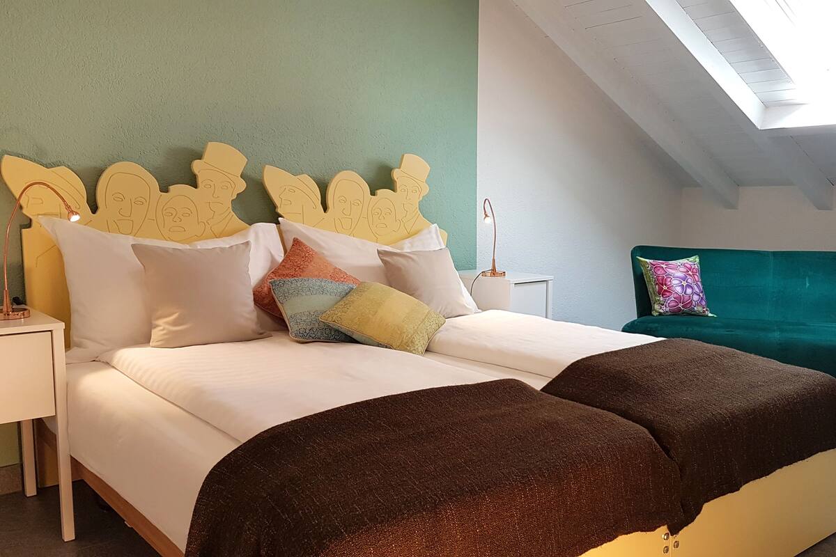 Un dormitorio con cama, almohadas, mesita de noche, escritorio, lámpara de escritorio, sofá verde y tragaluz.